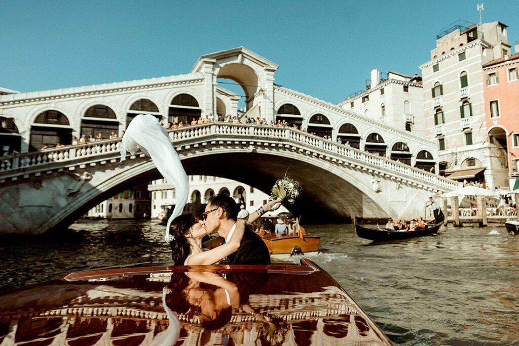 A wedding couple kissing on a boat under the Rialto Bridge in Venice with the bride's veil floating