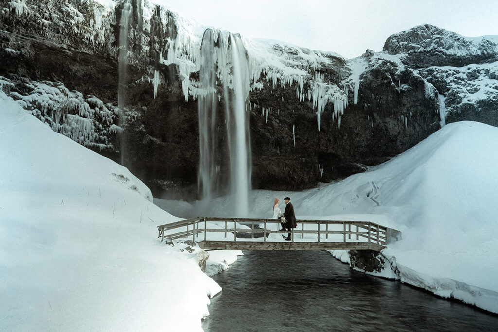Wedding couple standing on a bridge in the snow near a waterfallar