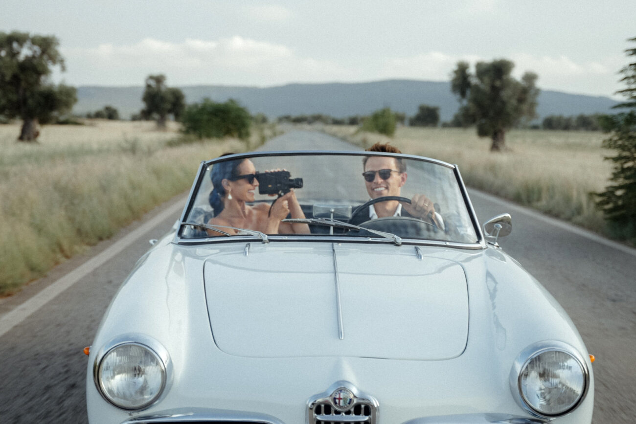 A bride films her groom in Super 8, while driving a white Alfa Romeo Spider after their Castello Marchione Wedding in Puglia