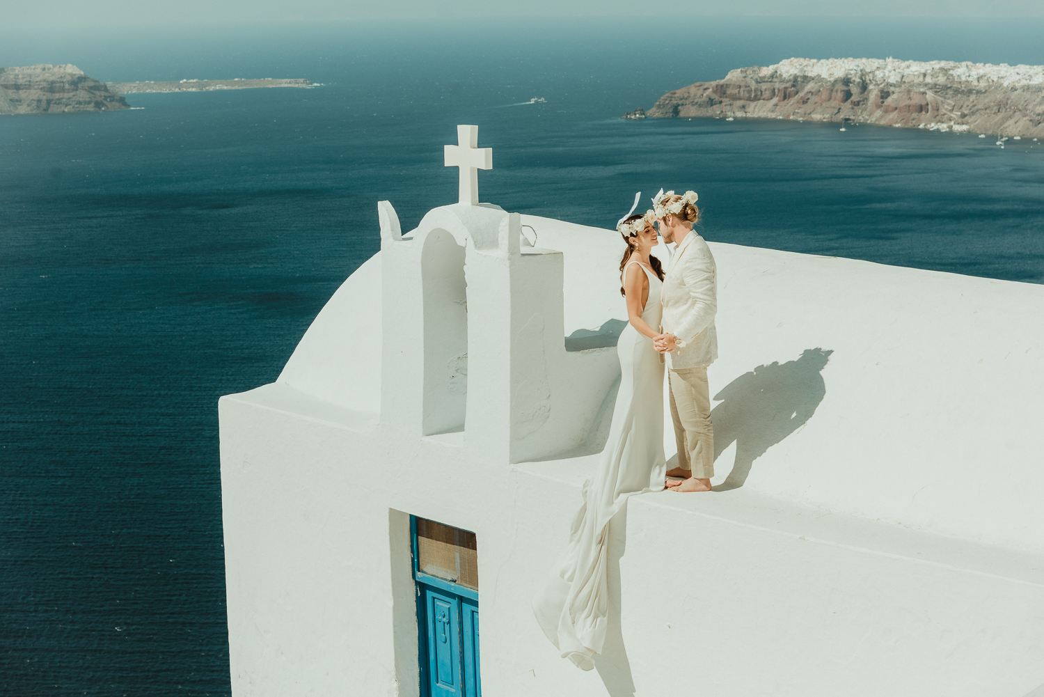 Couple on top of a white church in Santorini wearing flower crowns filmed by an Elopement Videographer for Intentional and Intimate Celebrations