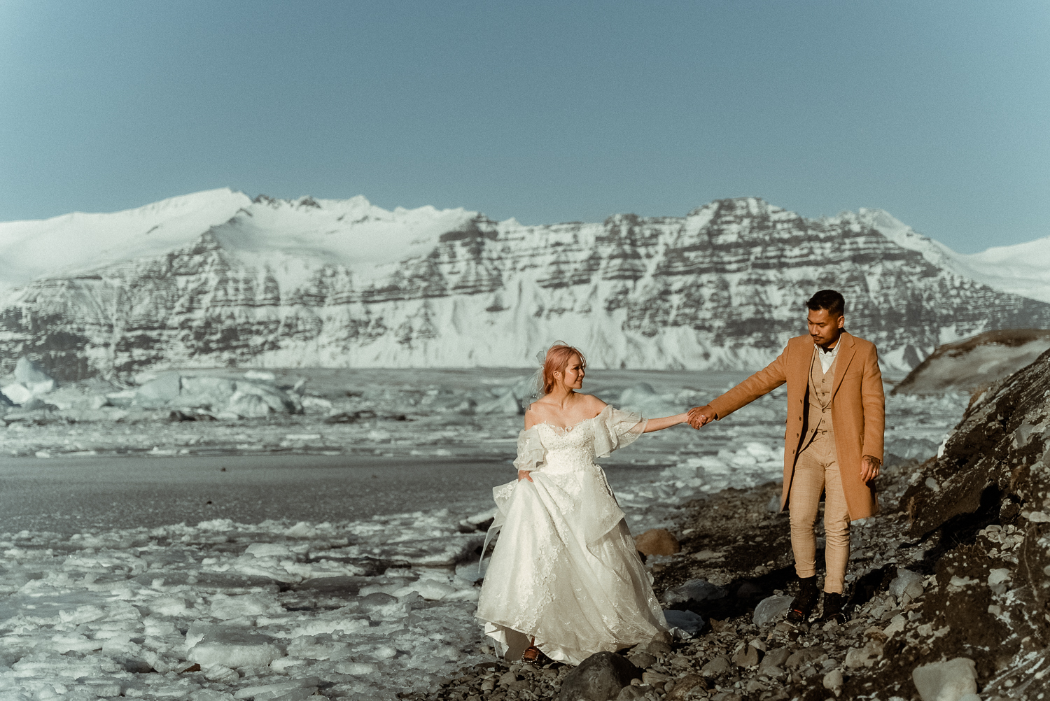 Iceland Elopement Videographer for Adventure & Intentional Celebrations 6 A couple in wedding attire walking hand in hand at the glacier lagoon, filmed by an Iceland Elopement Videographer
