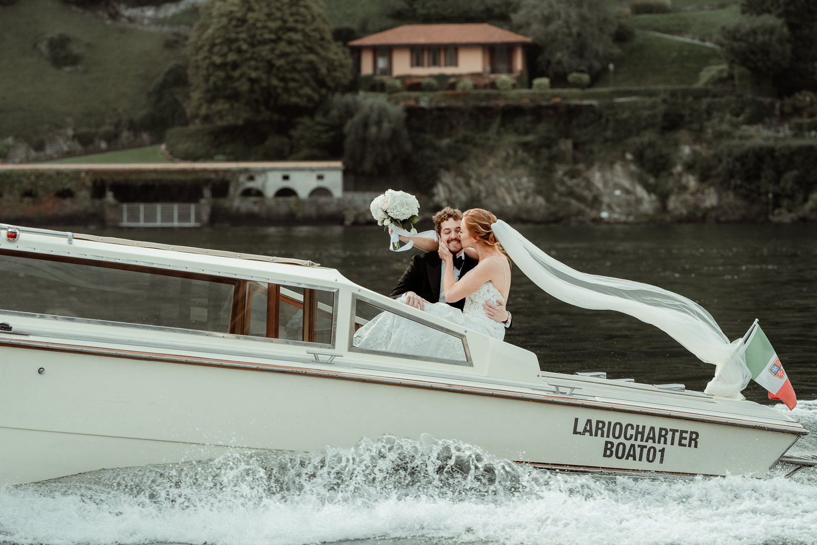 Bride kissing her groom on a boat in motion with her veil floating after their Intimate Lake Como Elopement at Villa Monastero