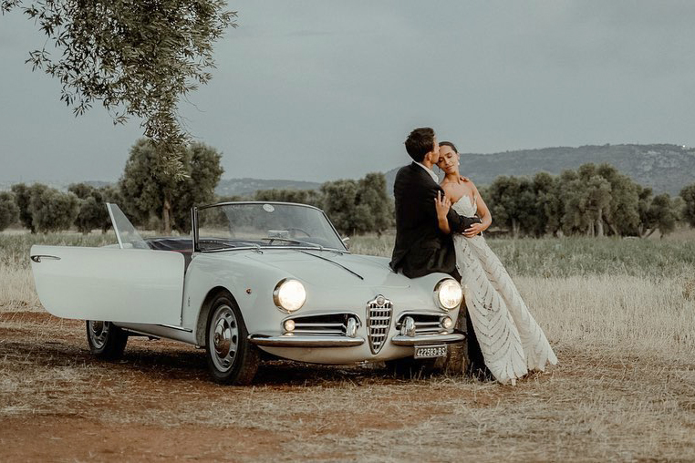 A couple in wedding attire sitting on a vintage Alfa Romeo Spider in a field of olive trees, filmed by a Puglia Wedding Videographer, after their Wedding at Castello Marchione