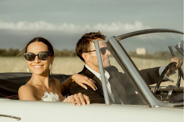 A couple in wedding attire, driving an Alfa Romeo Spider and wearing sunglasses after their Castello Marchione wedding in Puglia