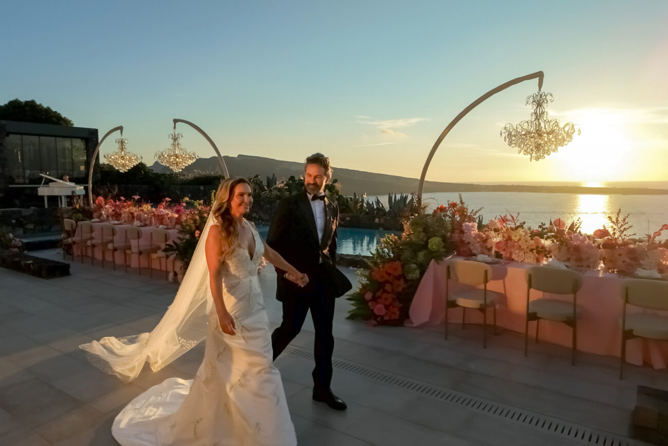 How to Plan a Destination Wedding in Greece, What to Know Before You Begin 9 wedding couple walking in Canaves Epitome at sunset in front of their dinner setup in a rose color decor from table tops to flowers and elegant chandeliers