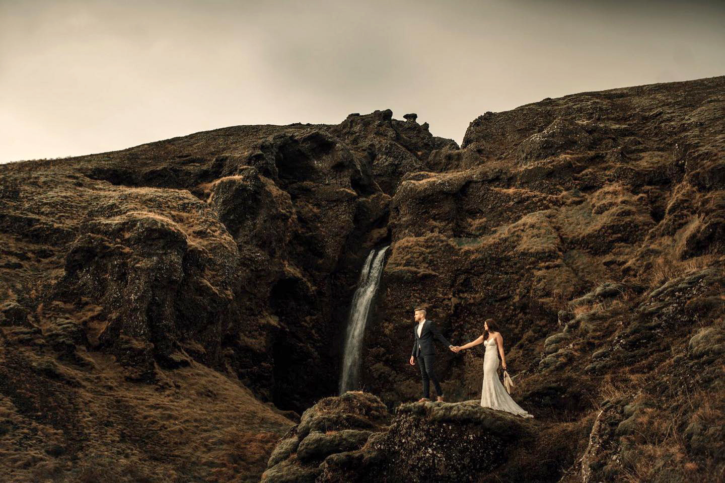 Iceland Elopement Videographer for Adventure & Intentional Celebrations 5 A couple in wedding attire walking uphill, with a secret waterfall visible in the background, filmed by an Iceland Elopement Videographer