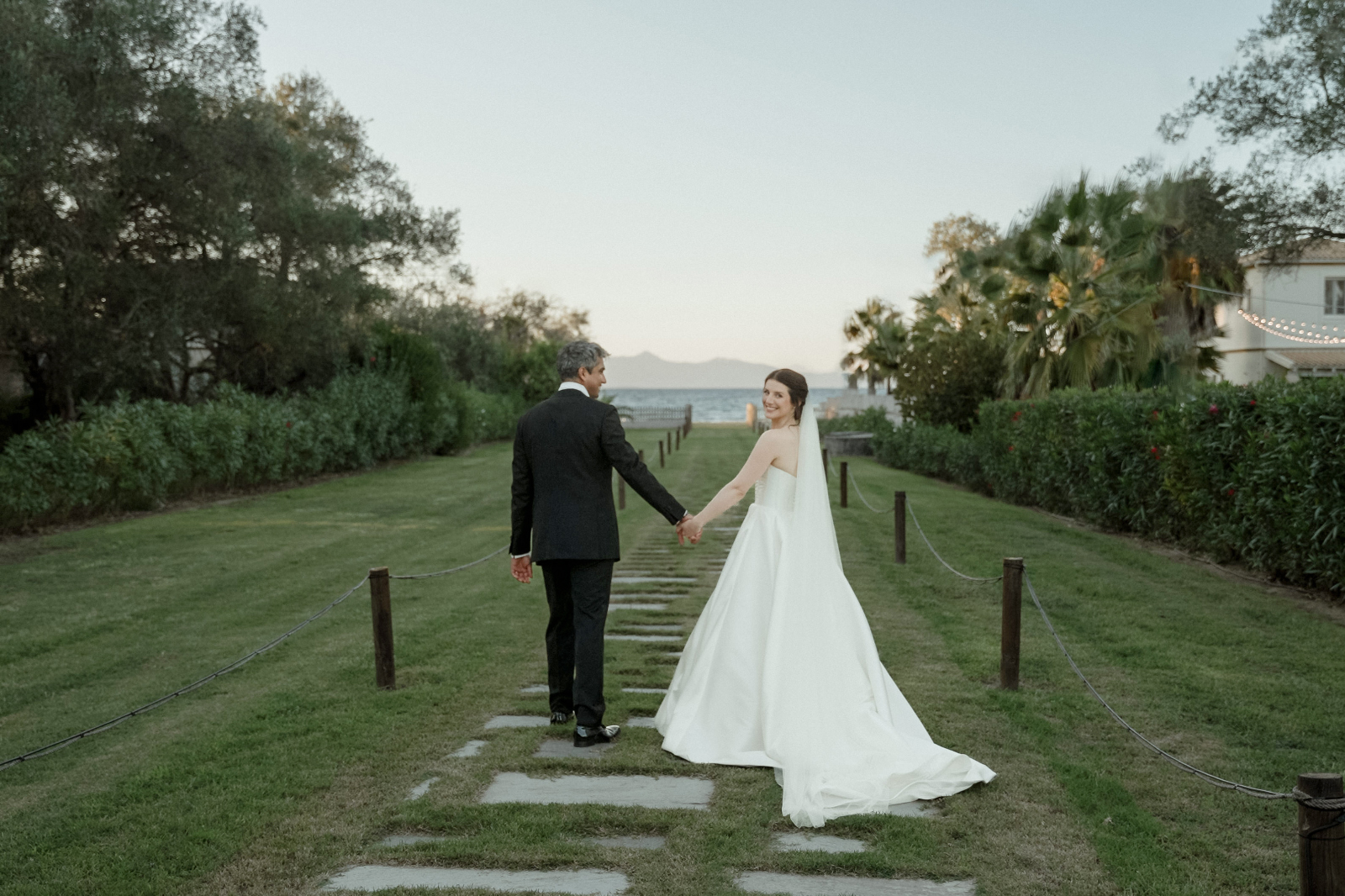 Indian Destination Wedding in Corfu at Natura Estate 10 A couple in wedding attire walks towards the beach and the bride looks back, after the first day of an Indian Destination Wedding in Corfu at Natura Estate