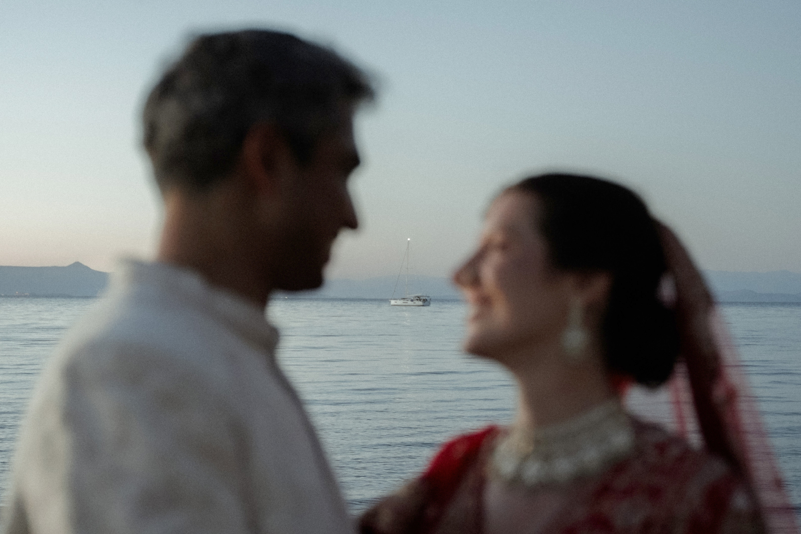 Indian Destination Wedding in Corfu at Natura Estate 7 A couple in indian wedding attire looking at each other on the beach at dusk, and a boat on the horizon in focus filmed for a destination wedding in Corfu at Natura Estate
