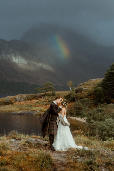 Couple kissing in wedding attire in front of a lake with a rainbow visible in the background in the Scottish Highlands, filmed by an elopement videographer