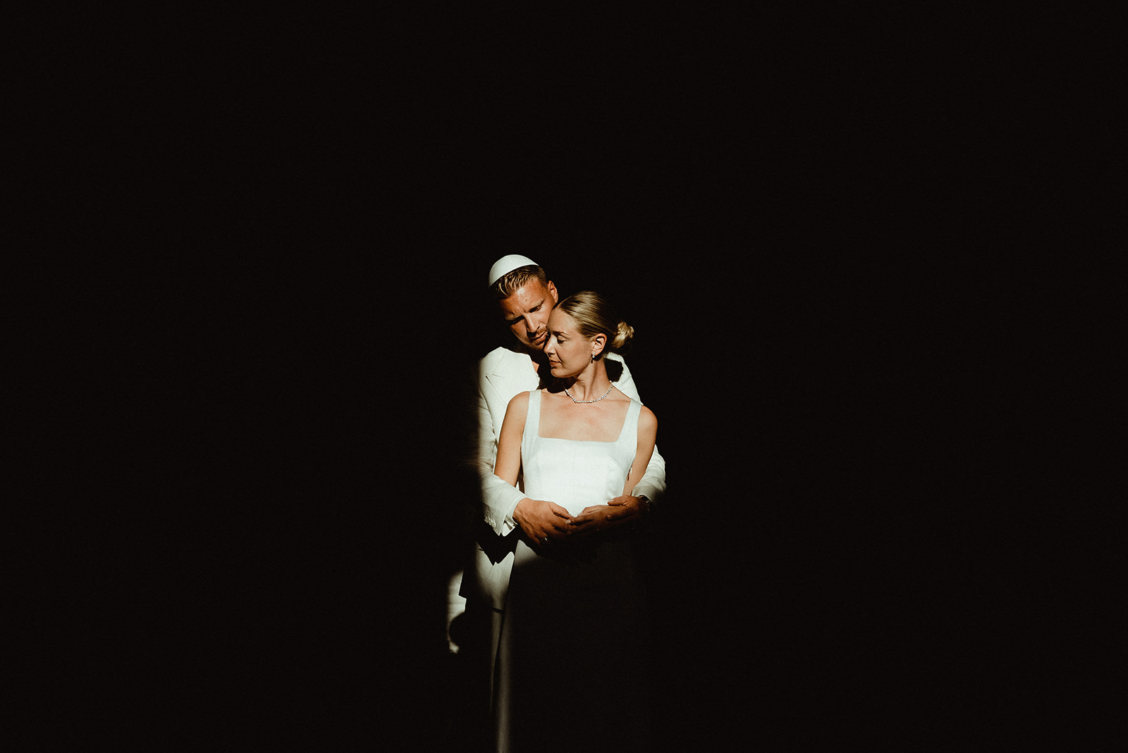 A couple dressed in white lit by a ray of sunlight, in the center of a dark frame before their Jewish Wedding in Morocco