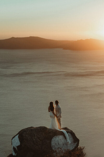 A couple standing on a rock above the sea gazing at the sunset in Imerovigli filmed by a Santorini Wedding Videographer