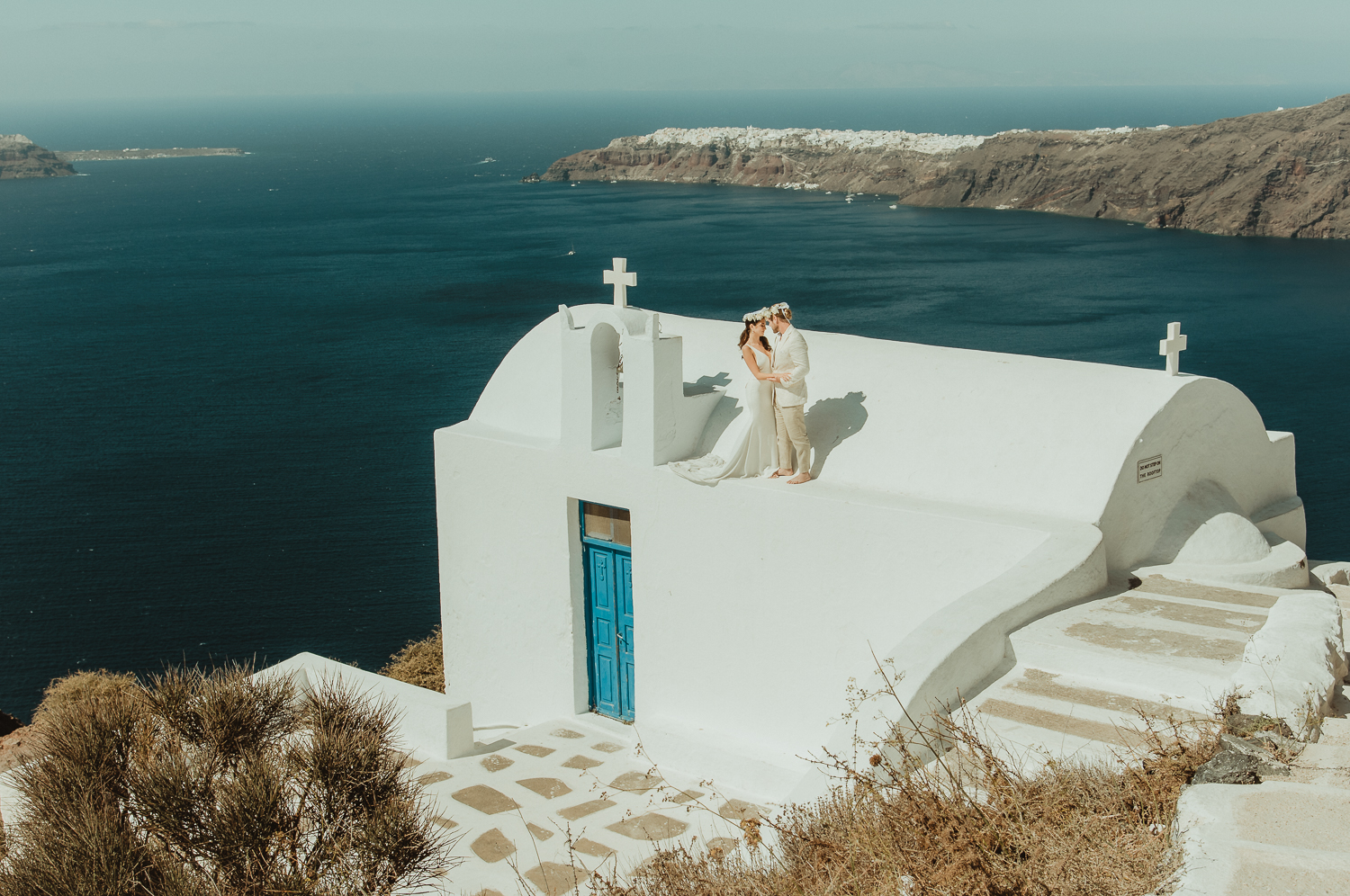 A wedding couple standing on the rooftop of a white church in Santorini wearing flower crowns illustration of ideas on how to plan a destination wedding in Greece