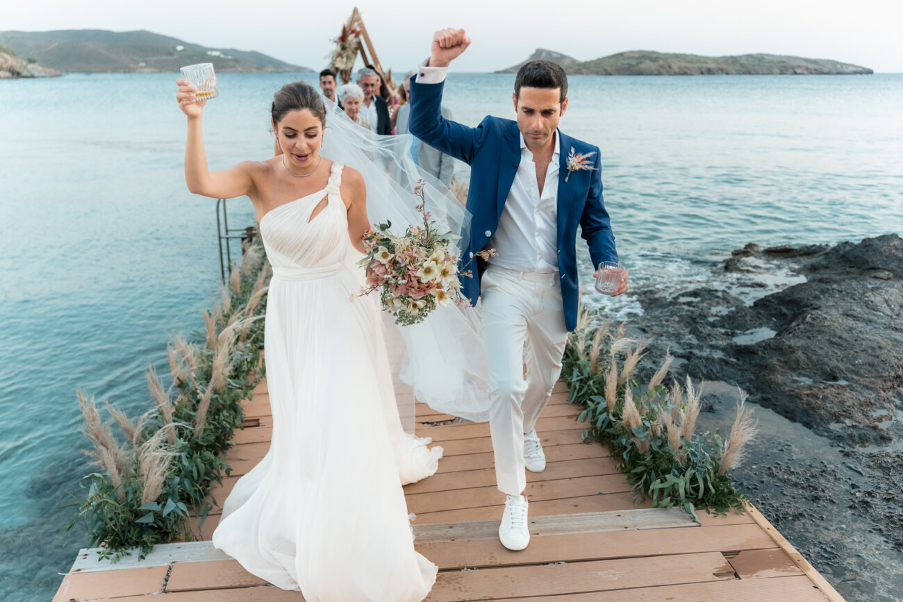 How to Plan a Destination Wedding in Greece, What to Know Before You Begin 1 A wedding couple cheering after their ceremony, walking on a deck by the sea in Syros, as a set of ideas on how to plan a destination wedding in Greece
