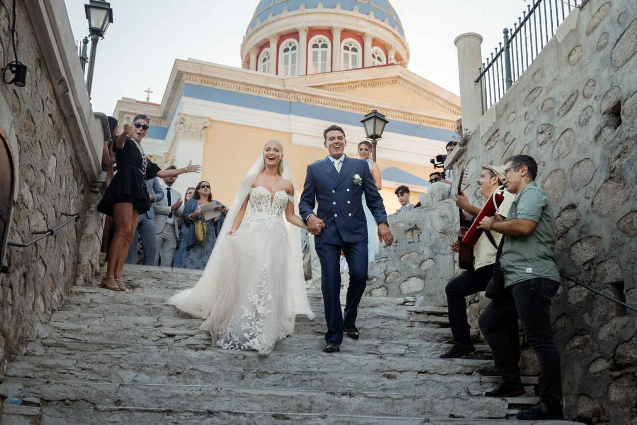 How to Plan a Destination Wedding in Greece, What to Know Before You Begin 14 A wedding couple walking down the steps in Syros, smiling while musicians play music for the couple, as a set of ideas on how to plan a destination wedding in Greece