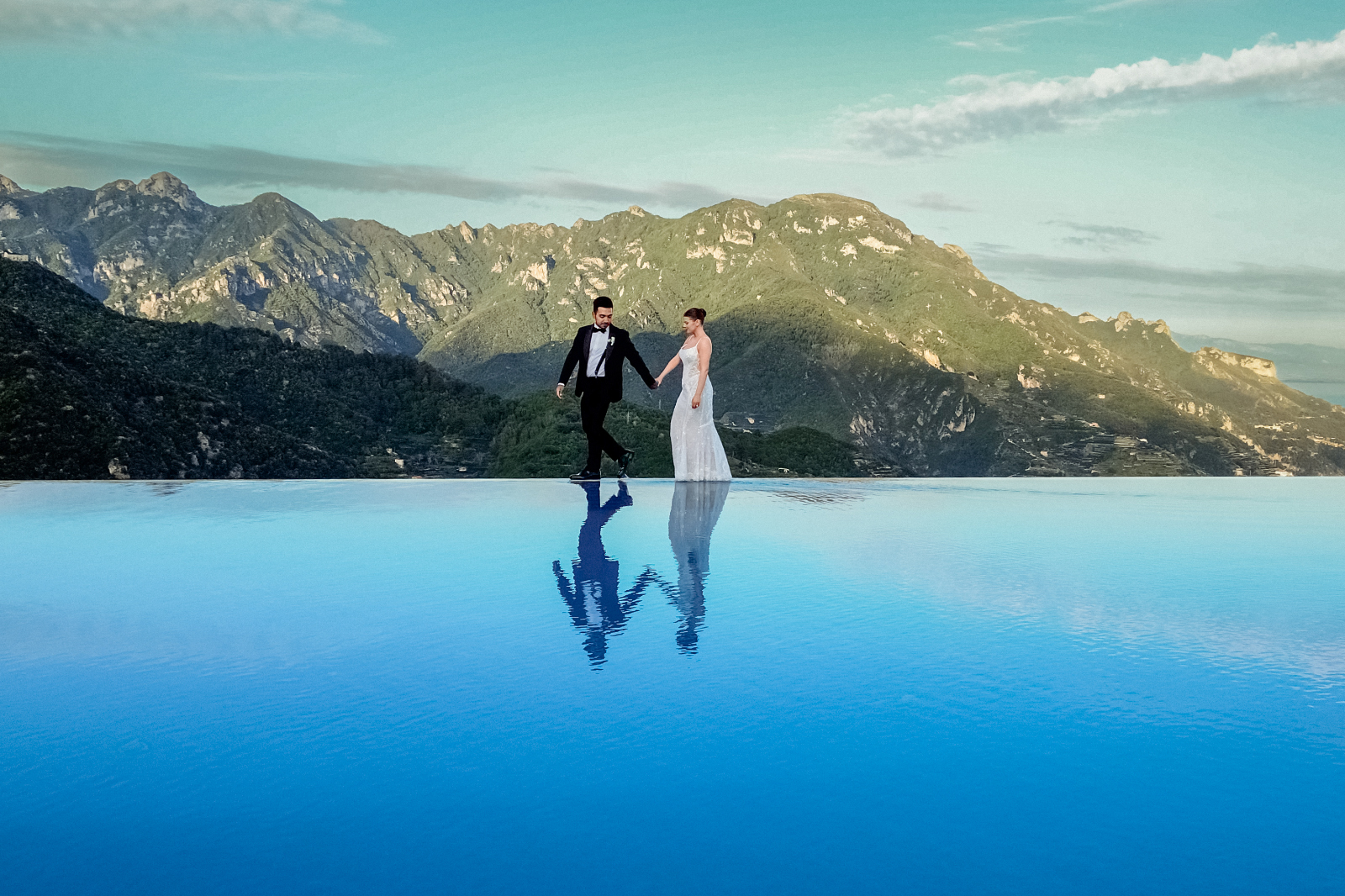 Bride and groom walking on the infinity pool with a stunning mountain background after their Belmond Caruso Wedding in Ravello
